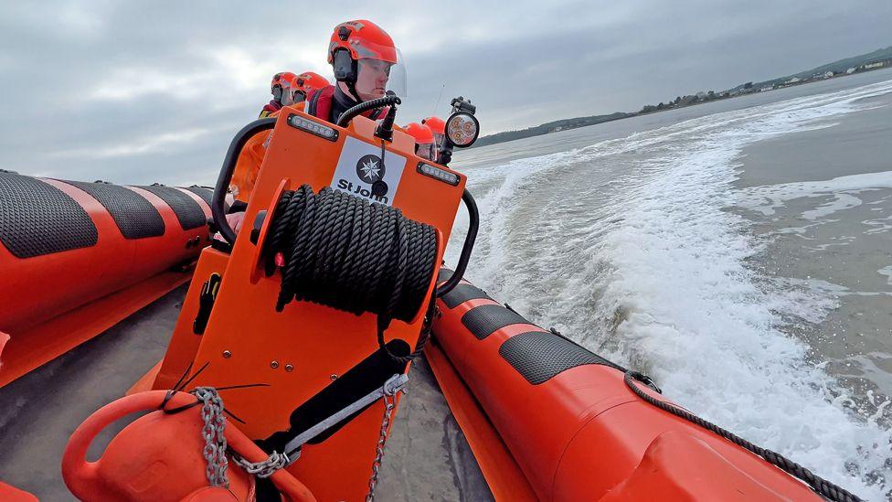 A man at the helm of an orange lifeboat steers it quickly through the waters leaving a big wake in its trail