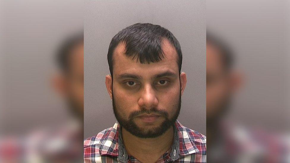 Police custody image of Mohammed Ali he has short dark hair and beard and a checked shirt