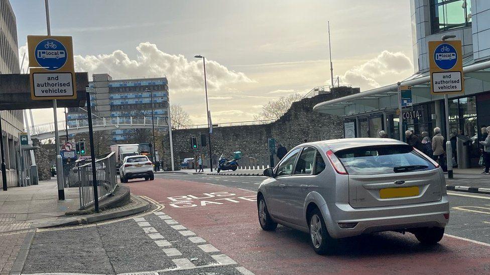 A silver car passes bus gate signs at Portland Terrace in Southampton. Ahead of it, a white car is also in the pink road lane, which is marked "bus gate" in white lettering.