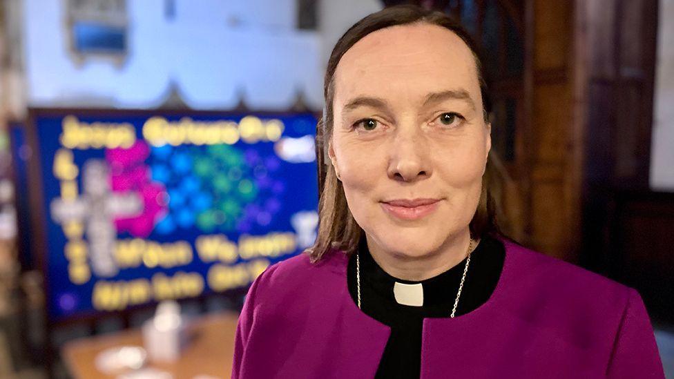 The Bishop of Hull, The Right Reverend Dr Eleanor Sanderson she has dark brown shoulder-length hair and is wearing purple and black vestments and a white clerical collar.