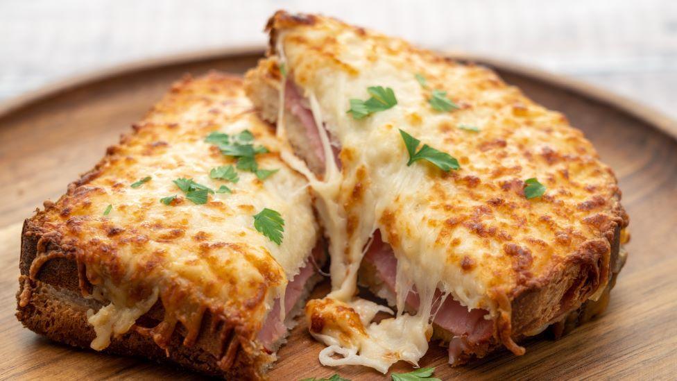 A croque monsieur, a sandwich with toasted bread, ham, and melted cheese on top with parsley, in served on wooden plate