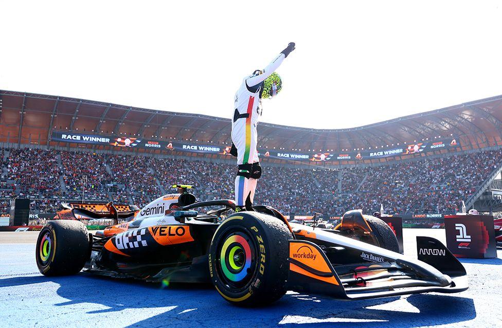 A race car driver in a white racing suit stands on top of a black and orange Formula 1 car with colourful wheel rims, raising one arm in celebration. The car is on a blue track surface, with a packed grandstand and “Race Winner” banners in the background.