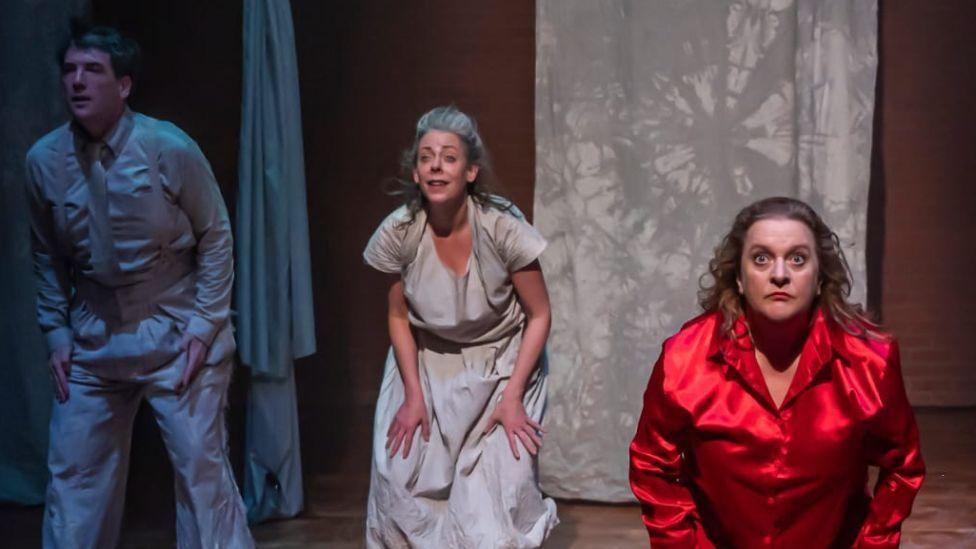 Still photography from Faust-ish play - a woman with blonde hair, in red silk pyjamas, stairs intensely to the audience. Beside her a man and woman, both in grey bedwear, look slightly to the side. The staging is minimal, with several sheets draped across a red brick background.