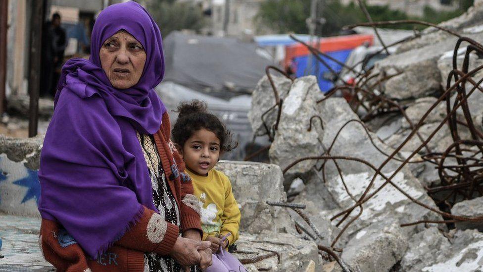 A woman wearing a purple shawl and a child sit next to a ruined building in Rafah (January 2024)