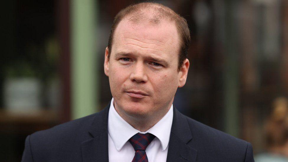 Gordon Lyons who is balding with light brown hair is wearing a dark suit, white shirt and black and red tie