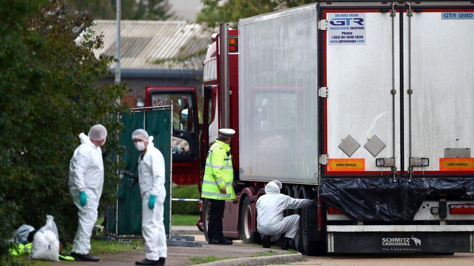 There is a large white and red lorry by the side of a road. There is a police officer nearby wearing a yellow jacket and black trousers and three forensic people in white suits, masks and black boots. One forensic person is kneeling down and reaching into a tyre area of the lorry. 