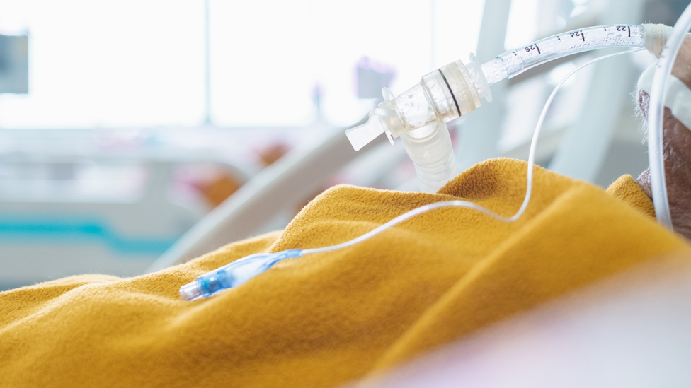 A close up of a person in a hospital bed with a yellow blanket over their body, and a tube in their mouth.