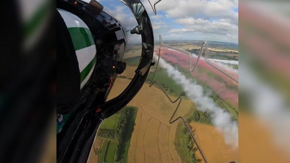 An aerial view of yellow and green fields beneath a plane, with white and red jets of stream. 