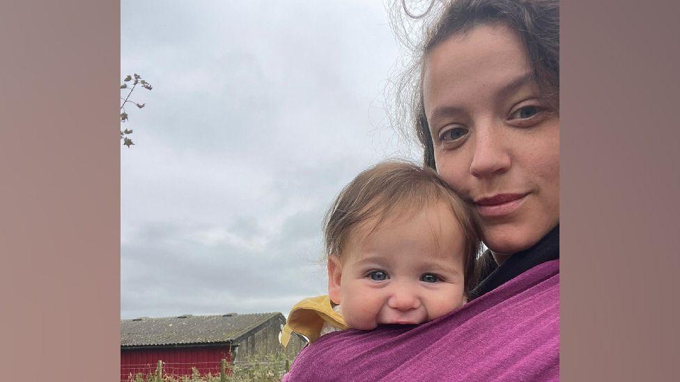 Mother Hannah Howell (right) has brown hair and blue eyes and looks into the camera. While her 7-month-old baby with blue eyes and brown hair strapped to her chest smiles into the camera. 