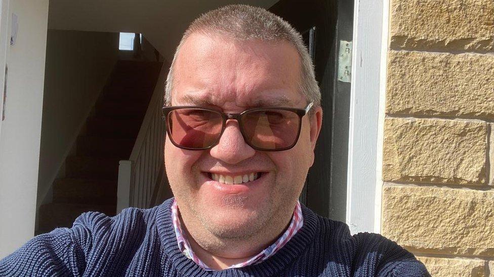 Chris has short cropped grey hair and is wearing sunglasses on his face and a blue jumper over a checked shirt. He is standing in front of a house while taking this selfie. 