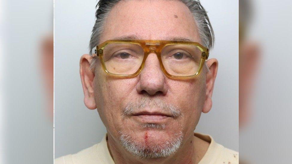 A police custody photo of a man with grey hair and grey stubble, wearing a pair of yellow glasses and a pale T-shirt. He's looking straight at the camera and has a plain white wall behind him.