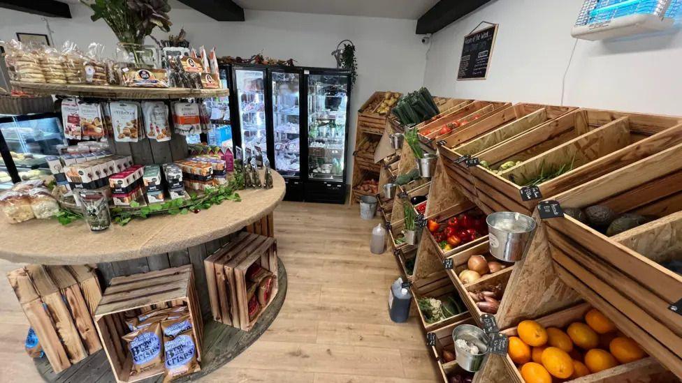 The Little Farm Shop in Shepton Mallet. There are crates with fruit, a table with chocolates and crisps, and a fridge in the background.