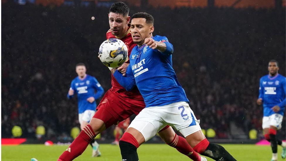 Two footballers, one in a red Aberdeen kit and the other in a blue Rangers kit, compete for a bouncing ball at Hampden 