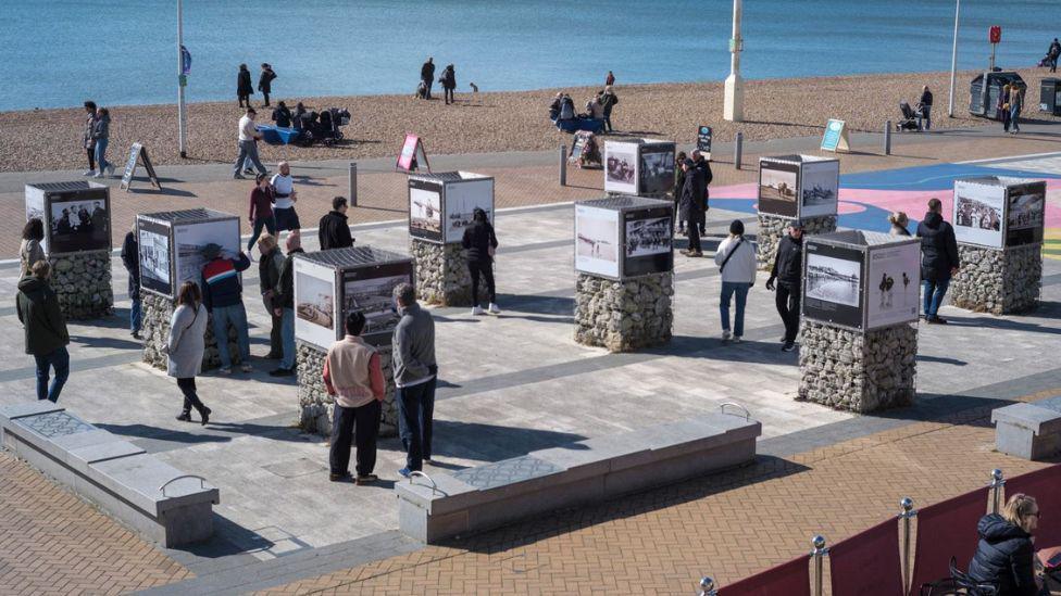 A dozen or so people are looking at a number of images. There is a pebbled beach in the background, as well as the sea. 