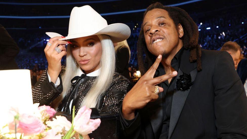 Jay-Z and Beyoncé pictured at the 2024 Grammy Awards. Beyonce is tipping a white cowboy hat, and Jay Z, wearing an all-black suit is gesturing to the camera with his hands.