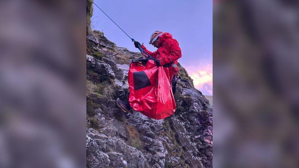 Man in helmet and red jacket holding a red bag while he climbs down a mountain attached to a rope.