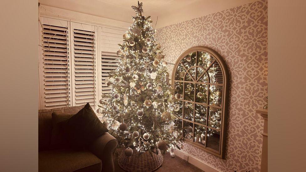 A Christmas tree is decorated with silver baubles and white lights in the corner of a living room next to a sofa and mirror.