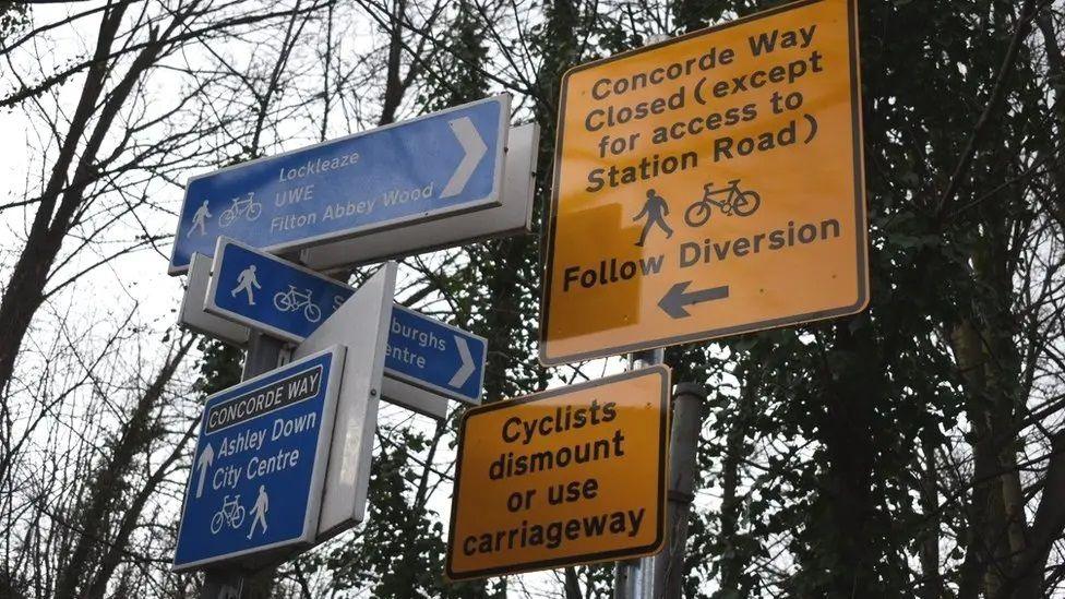 Road signs displayed in Lockleaze. There are three separate blue signs in the direction of Lockleaze, St Werburghs and Ashley Down. There are larger yellow signs which say that Concorde Way is closed and that cyclists should dismount or use the carriageway.