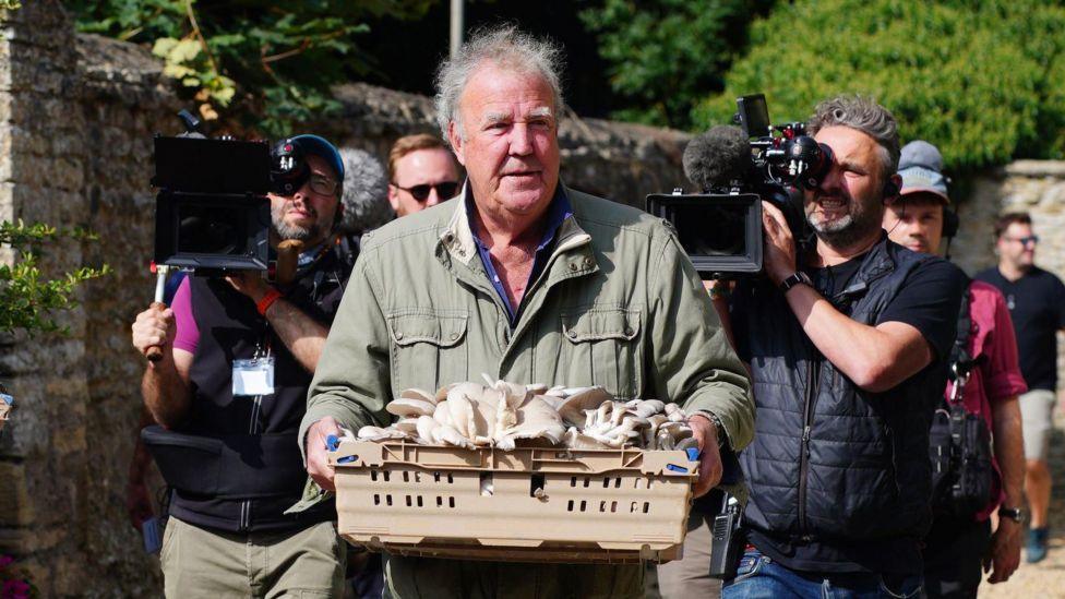 Photo of Jeremy Clarkson carrying a pallet of food. Behind him are two TV crews