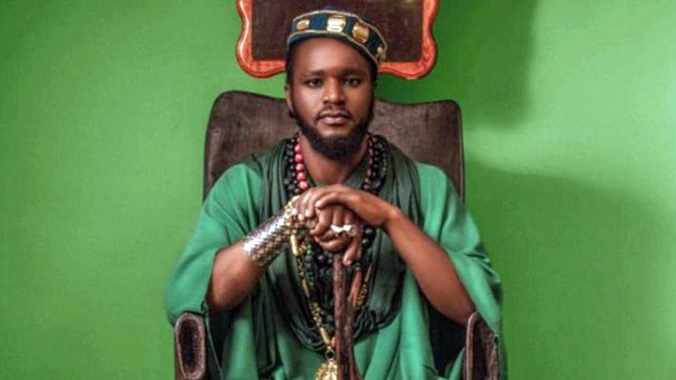 Aamron pictured sitting on a beautifully carved wooden chair wearing a green gown, decorated green hat, lots of beads and other jewellery. His hands rest on a ceremonial walking stick. Behind him the background is bright green.