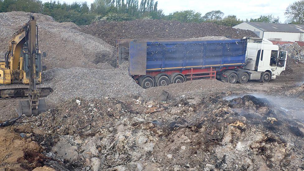 A lorry and digger at the site surrounded by piles of burnt waste 