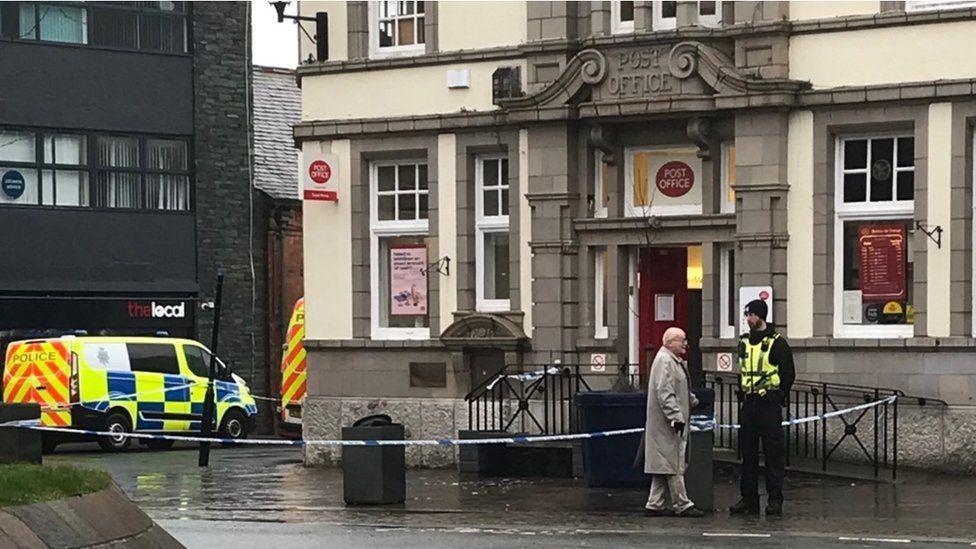 A police cordon around an area of pavement outside a post office in Ulverston, where Holly Lambert died. A police van is parked around the corner and an officer is standing watch by the police tape.