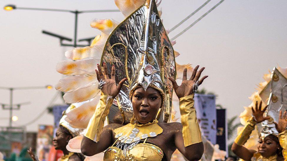 A performer in a gold outfit with gold glove sleeves and large silver headdress does jazz hands during the Calabar Carnival