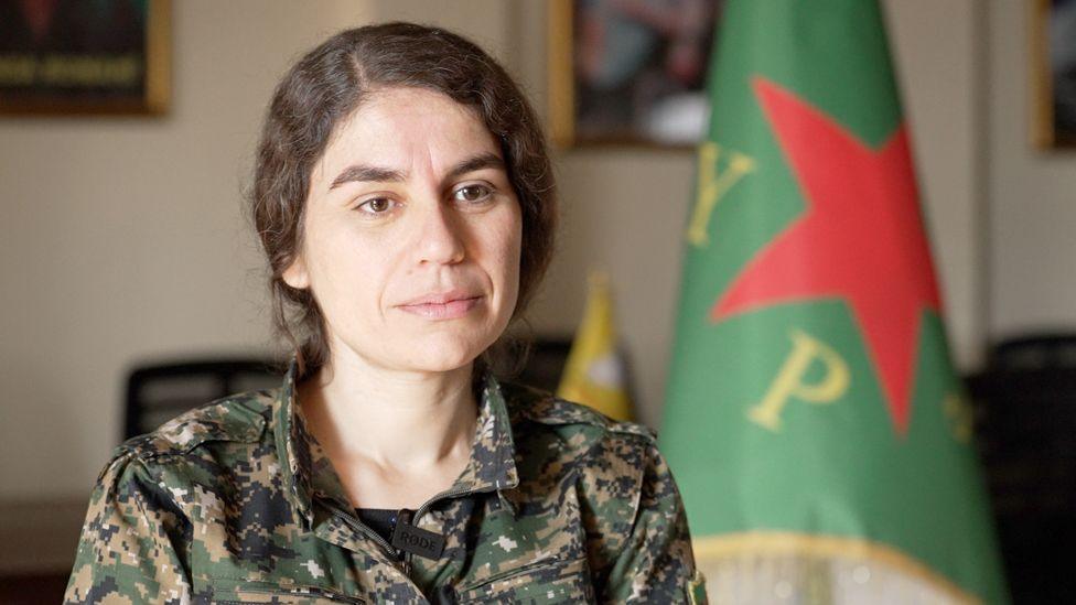 Headshot of Roksana Mohamed wearing army uniform - she has long dark hair which is tied back. A green flag with a red star and the letters Y and P can be seen behind her.