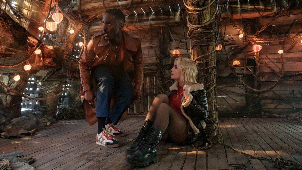 Ncuti Gatwa, as The Doctor, wearing a long brown coat and coloured sneakers, crouches down to talk to his assistant - a blonde woman in a leather jacket - who is tied to a pole inside a wooden cabin.