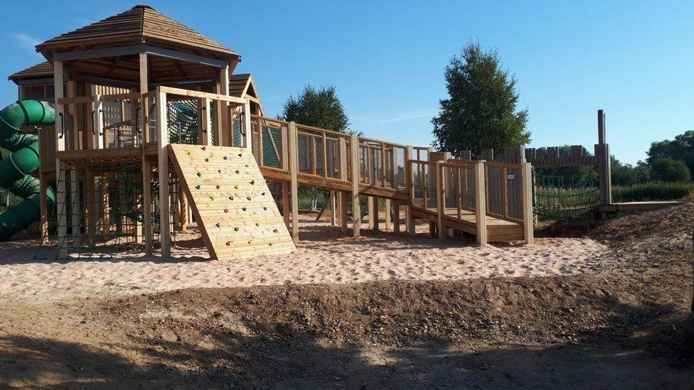 A wooden tower and ramp. The is a wooden climbing wall in front of the tower and there is sand and mud on the ground.