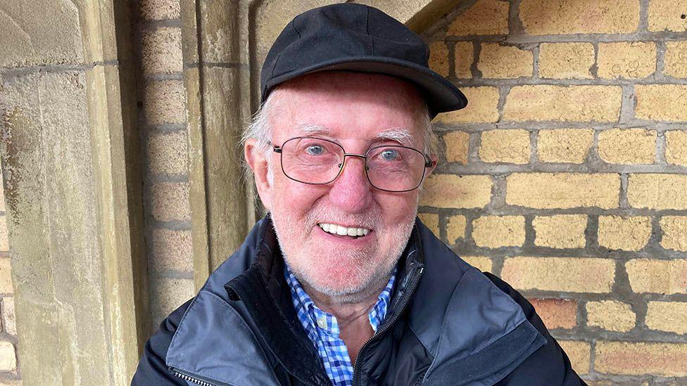 Tom Brett stands outside Preston railway station in front of a sandstone brick wall. He is wearing a black baseball cap and has short white hair, stubble and wire-framed glasses and a black coat.