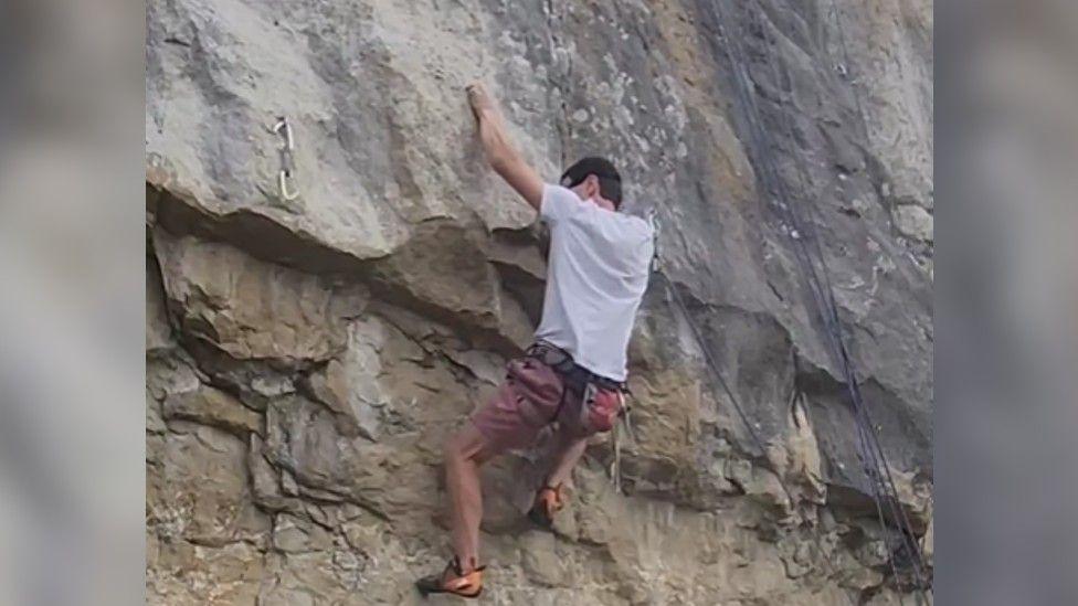 A man in a white t-shirt and pink shorts climbing a steep cliff face, secured to the rock with a harness