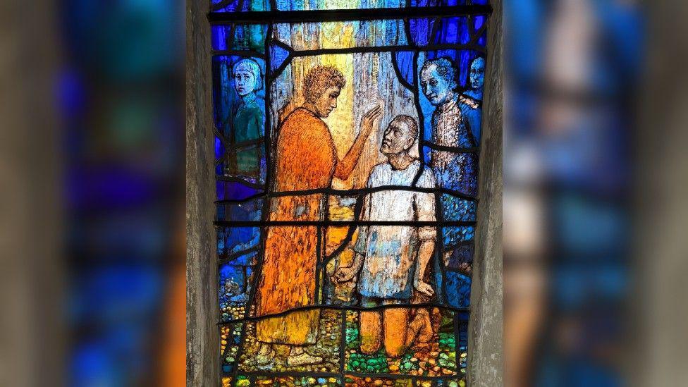 A close-up of a pane of stained glass with Jesus in an orange robe with his arm raised above a blind man who is kneeling before him