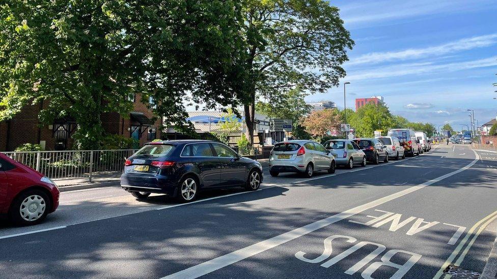 A section of Bitterne Road West shows a queue of cars waiting at a traffic lights, there are trees lining the left side of the road, with buildings behind the trees and a bus lane on the opposite side of the road