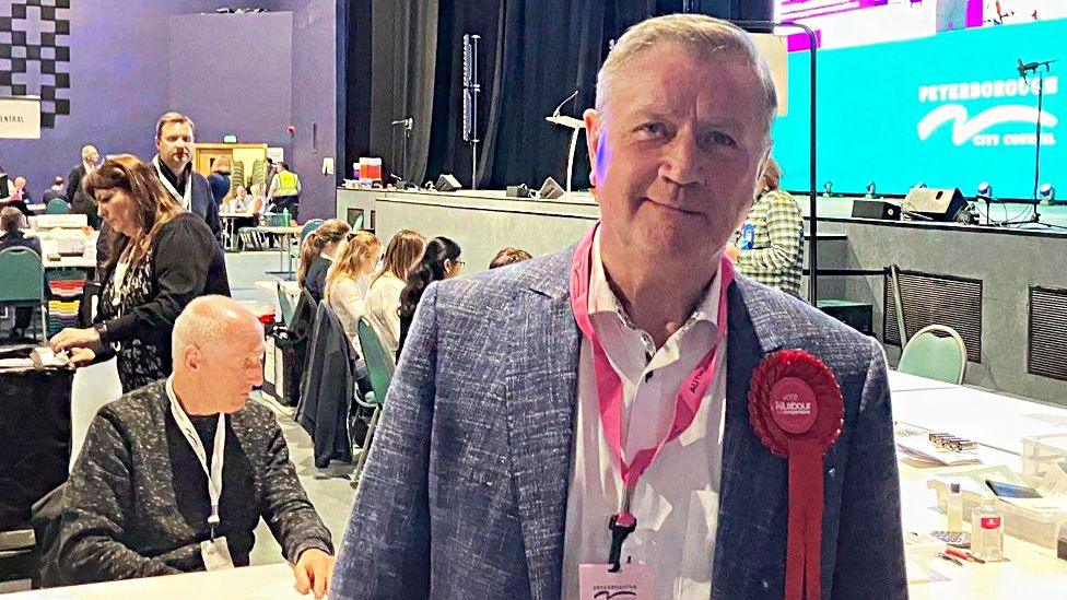 Dennis Jones at an election count, wearing a blue jacket with a red rosette. He is smiling for the camera.