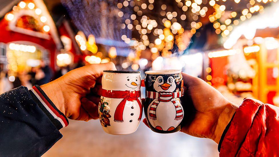 Two hands holding festive snowman and penguin mugs with the lights of stalls in the background