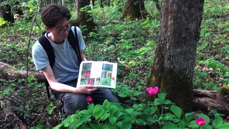 Kevin Lik squatting next to some pink flowers and holding a book identifying the plant