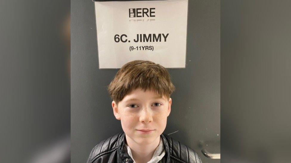 Albie Salter looking straight at the camera wearing a black leather jacket, standing in front of a black door with a white sheet of paper saying his character name "JIMMY".