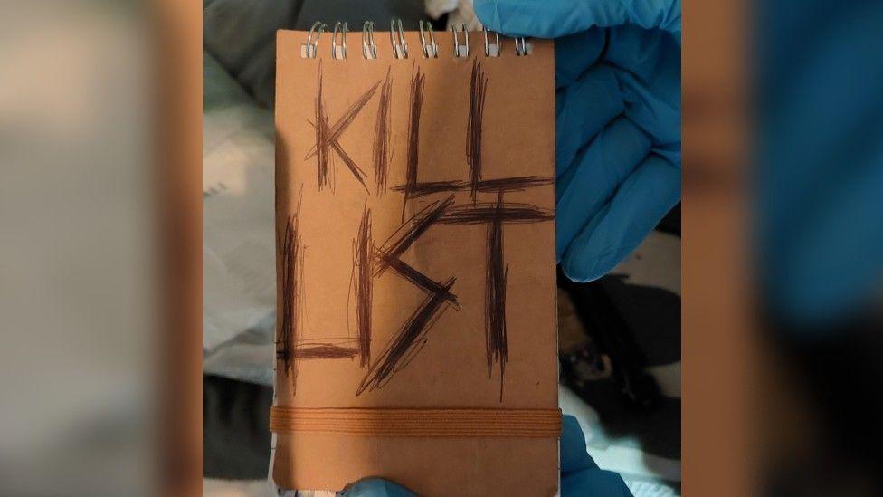 Latex gloved hands holding up a notebook with "kill list" written in capital letters on the front in black ink, drawn over many times.