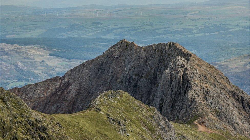 A steep mountain ridge, with a pathway in the forefront and mountain landscape behind.