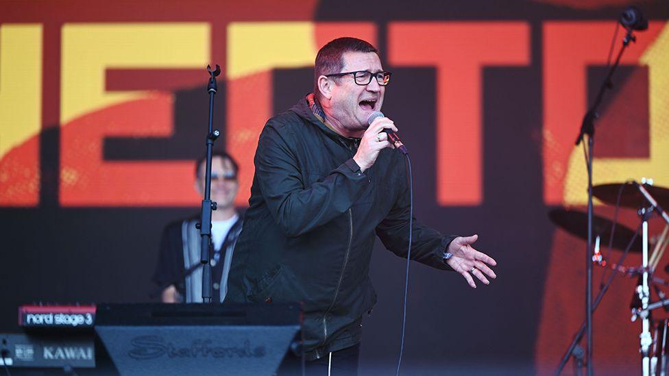 Paul Heaton in a dark jacket holding a microphone singing on stage at the Glastonbury Festival in 2024. He has short dark hair and is wearing black glasses.