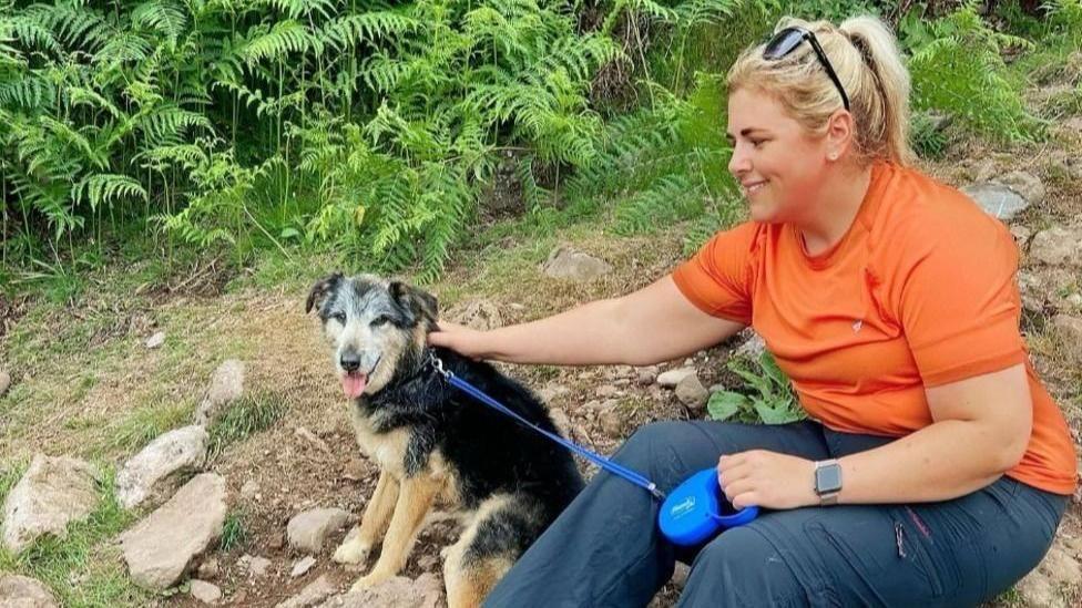 Adele Rennie, sitting on a rock outdoors, wearing an orange t-shirt and grey trousers. She has a dog on a lead and has blonde hair.