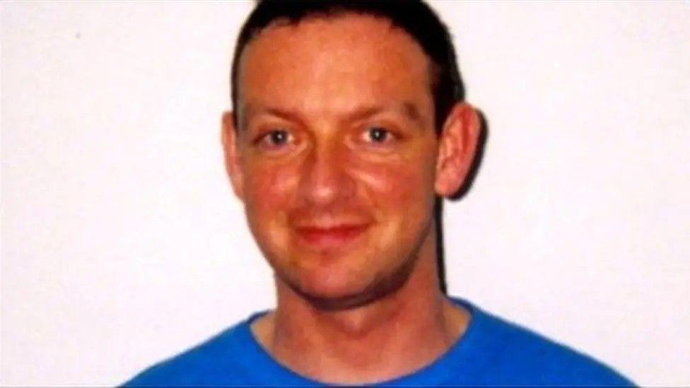 A man, with dark hair, smiling at the camera wearing a blue t shirt