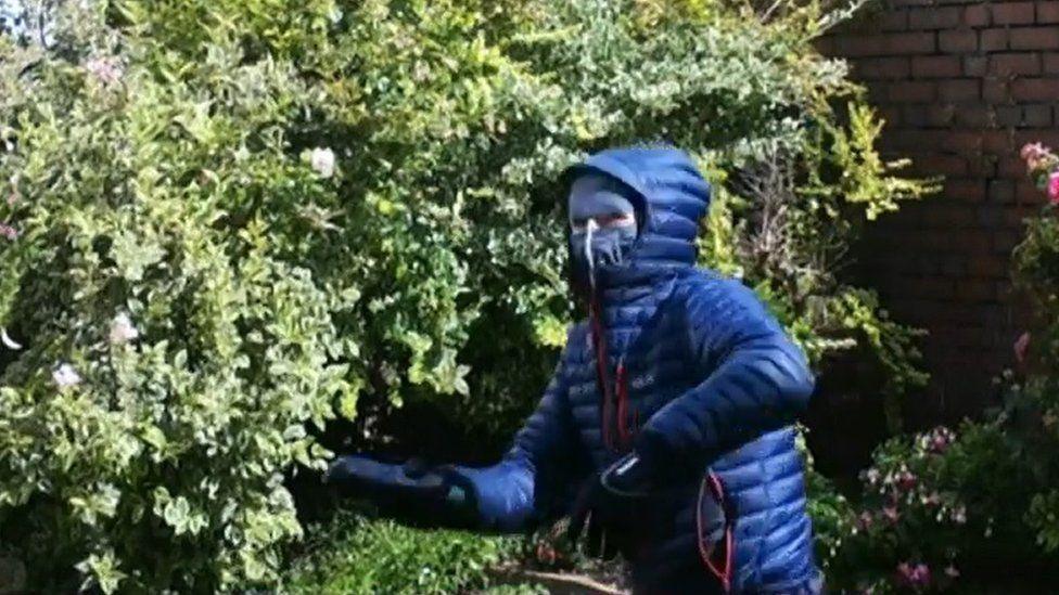 A person in a blue anorak, with the hood up and a mask over the face, starts to throw an unidentified package towards a green bush.