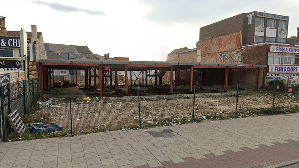 Derelict site of the hotel, showing a steel framework with rubbish and wood pallets behind a fence lining street