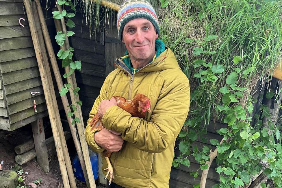Dave Jacobs smiling at camera and holding a chicken. He is wearing a mustard-coloured jacket and a multi-coloured wooly hat. There is greenery behind him and a wooden structure.