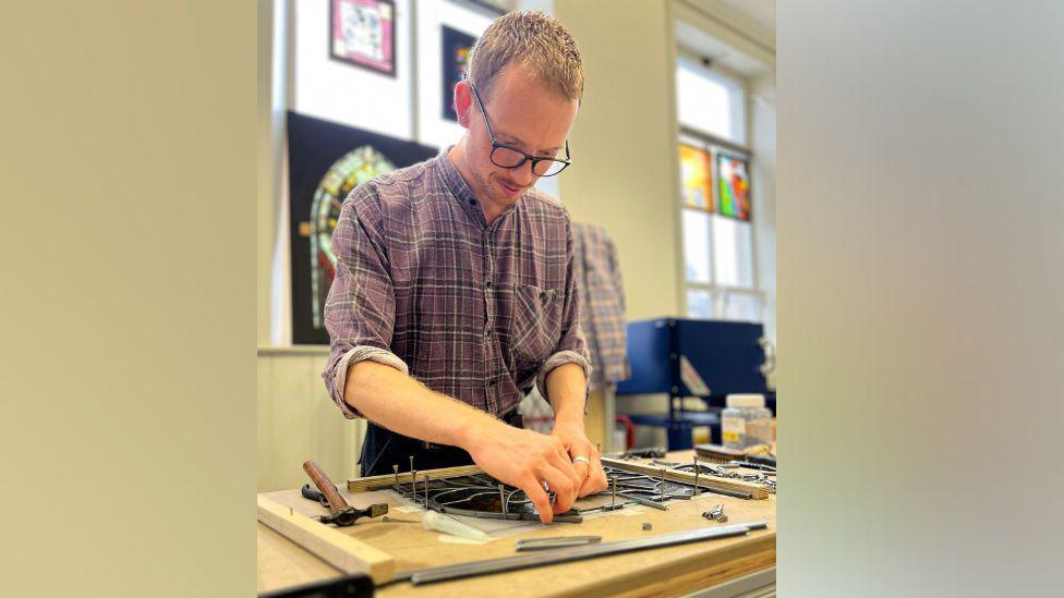 A man wearing a purple checked shirt and black-rimmed glasses is in a workshop restoring a panel of stained glass. The glass is on a wooden worktop with nails sticking out and is surrounded by tools including a hammer. In the background there are colourful glass panels propped up by the windows.