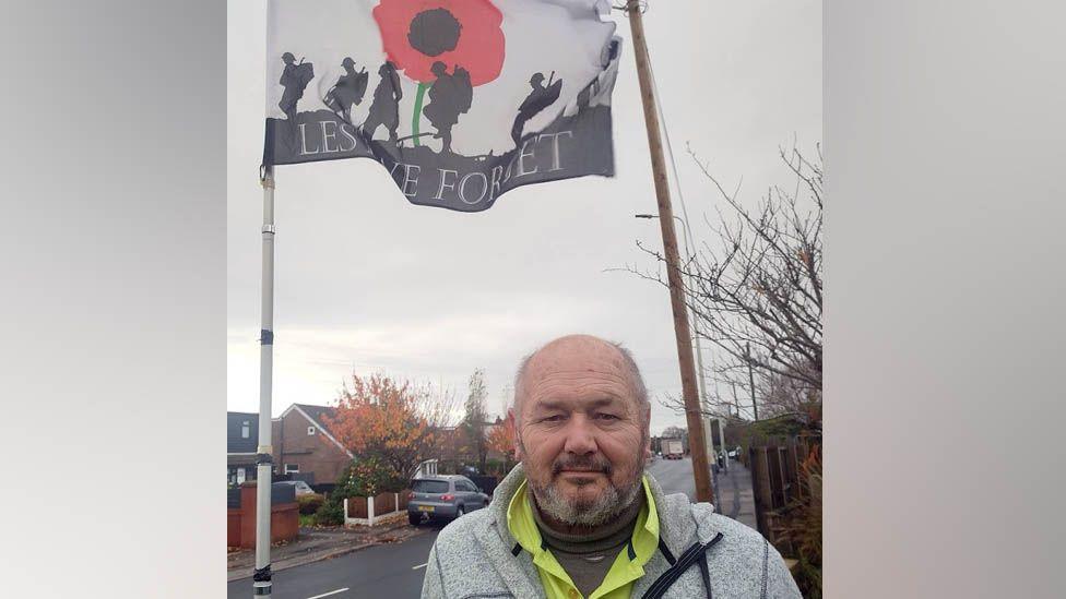 Steve Layton has short hair and is wearing a grey fleece and a yellow jacket. He stands in front of his flagpole which has outlines of solider with a red poppy with a road in the background