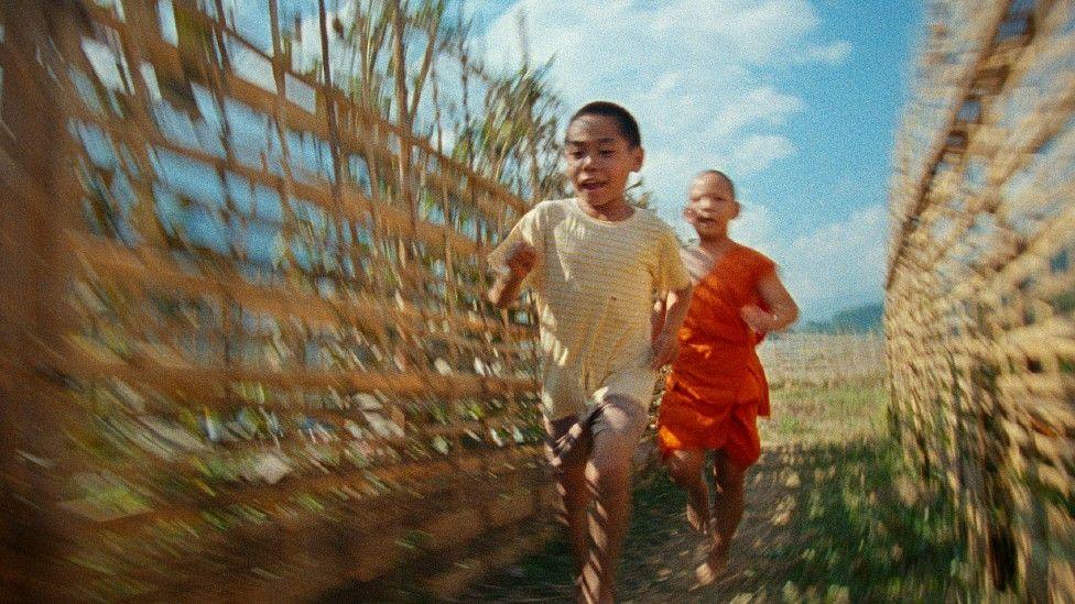 Blurred imaged suggesting motion of two Laotian children running between two fences with crops behind them