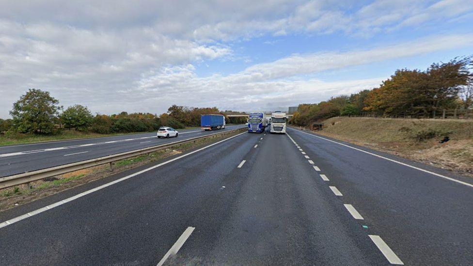 The M11 viewed from the right carriageway, with the central reservation. This is a google image from a different date so no lanes are closed. Two lorries side by side are coming towards the camera, while a white car and lorry are on the other side. 